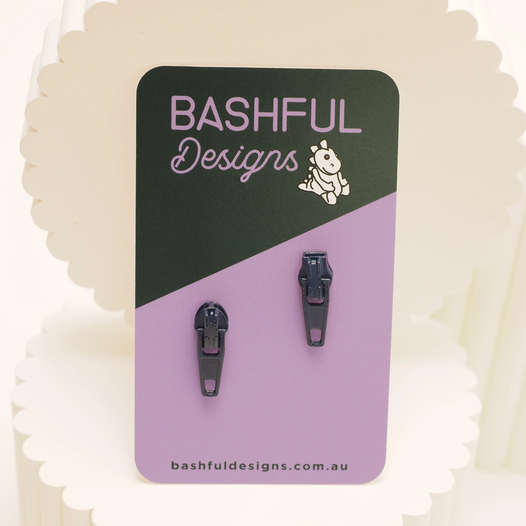 Dark blue zipper pull stud earrings on a 'Bashful Designs' branded card.
