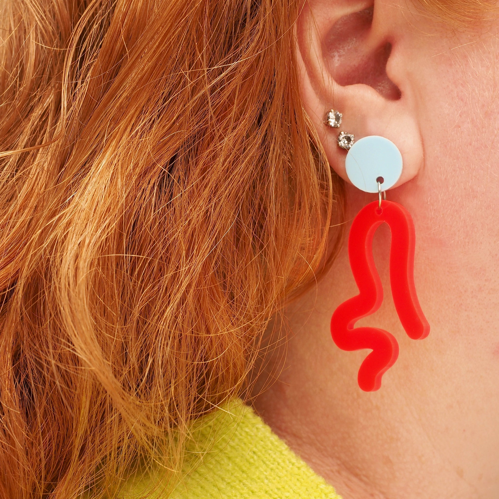 A person with red curly hair, wearing neon red acrylic wavy earrings, that look like a person twerking. These earrings have a light blue earring topper.