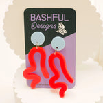 Neon red acrylic wavy earrings that look like a person twerking, on a 'Bashful Designs' branded card against a light background. These earrings have a light blue earring topper.