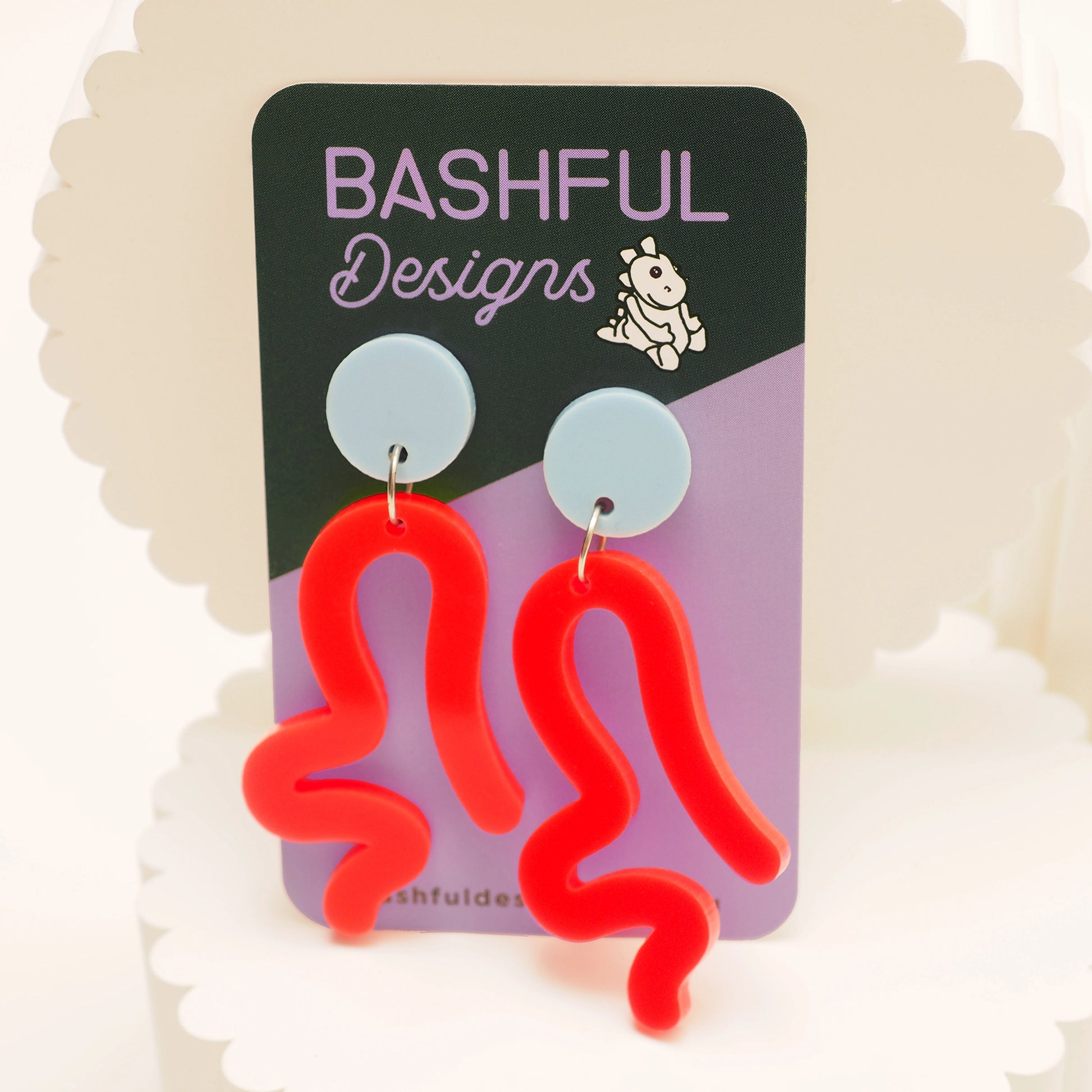 Neon red acrylic wavy earrings that look like a person twerking, on a 'Bashful Designs' branded card against a light background. These earrings have a light blue earring topper.