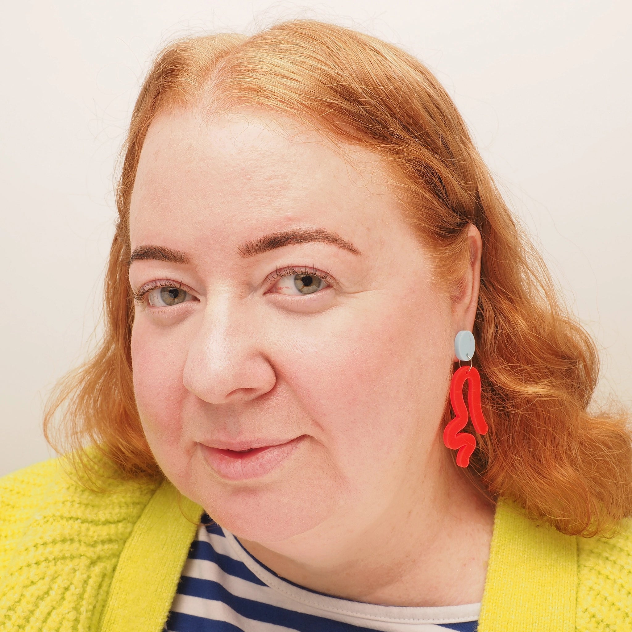 A woman with curly red hair, wearing neon red acrylic earrings, in the shape of a person twerking.