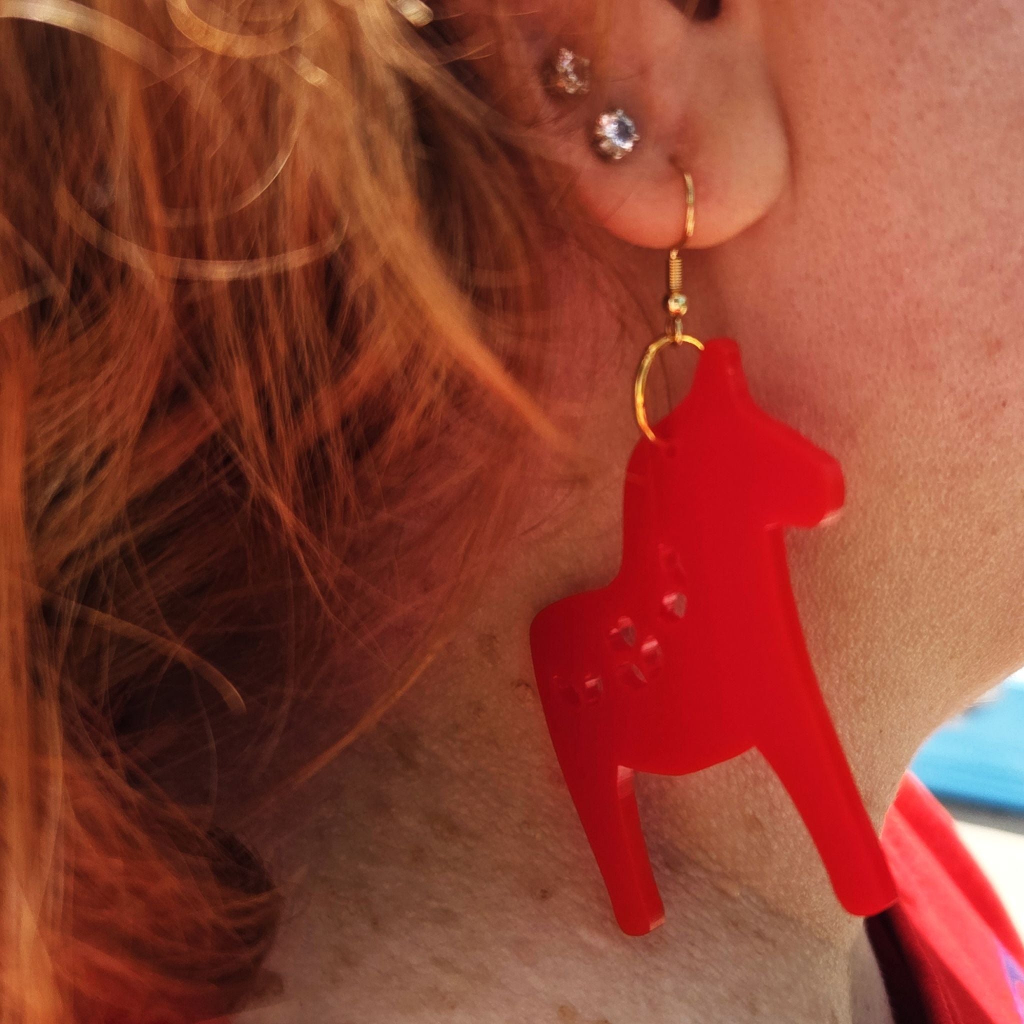 Earrings in a shape of a Nordic Christmas horse, made in red acrylic.
These are shown hanging in a person's ear.