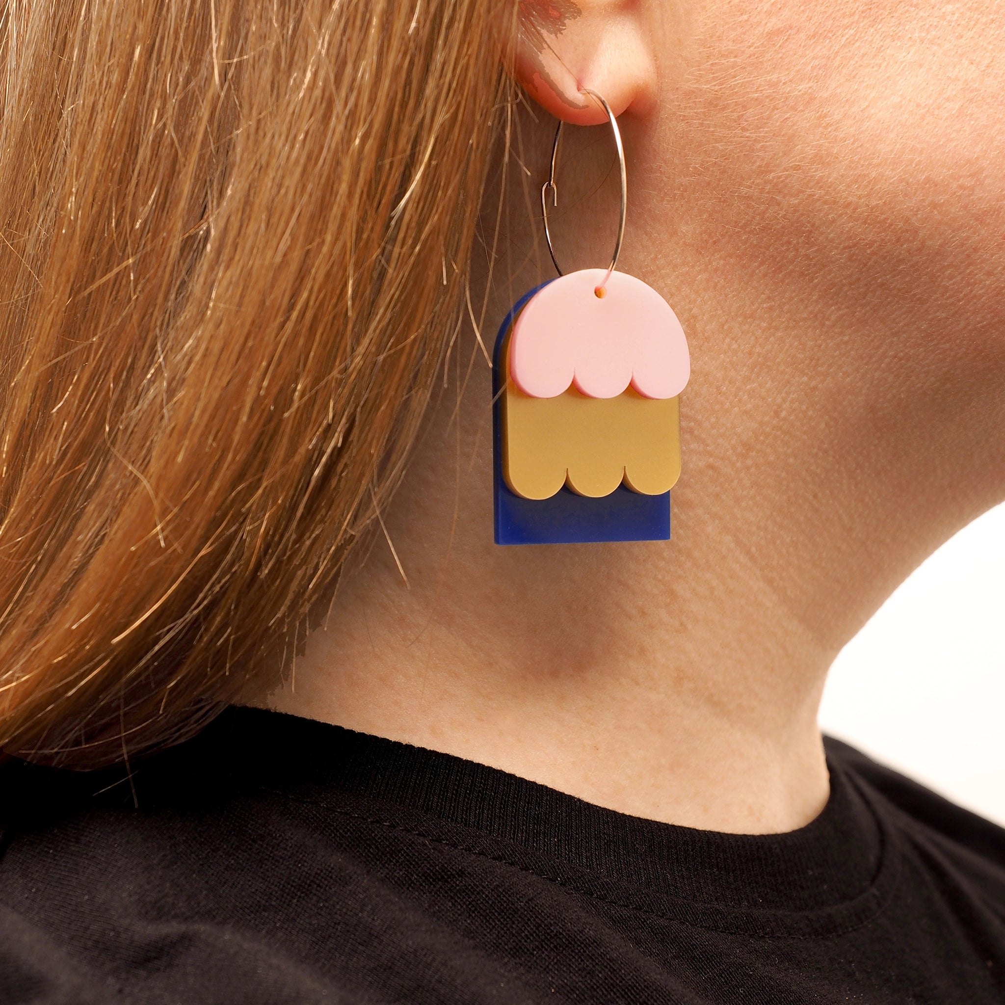 A person with straight red hair, wearing earrings shaped like layered petals. These earrings have three layers, one is blue, one if pink and one is gold. They are hanging on a large hoop fixture.