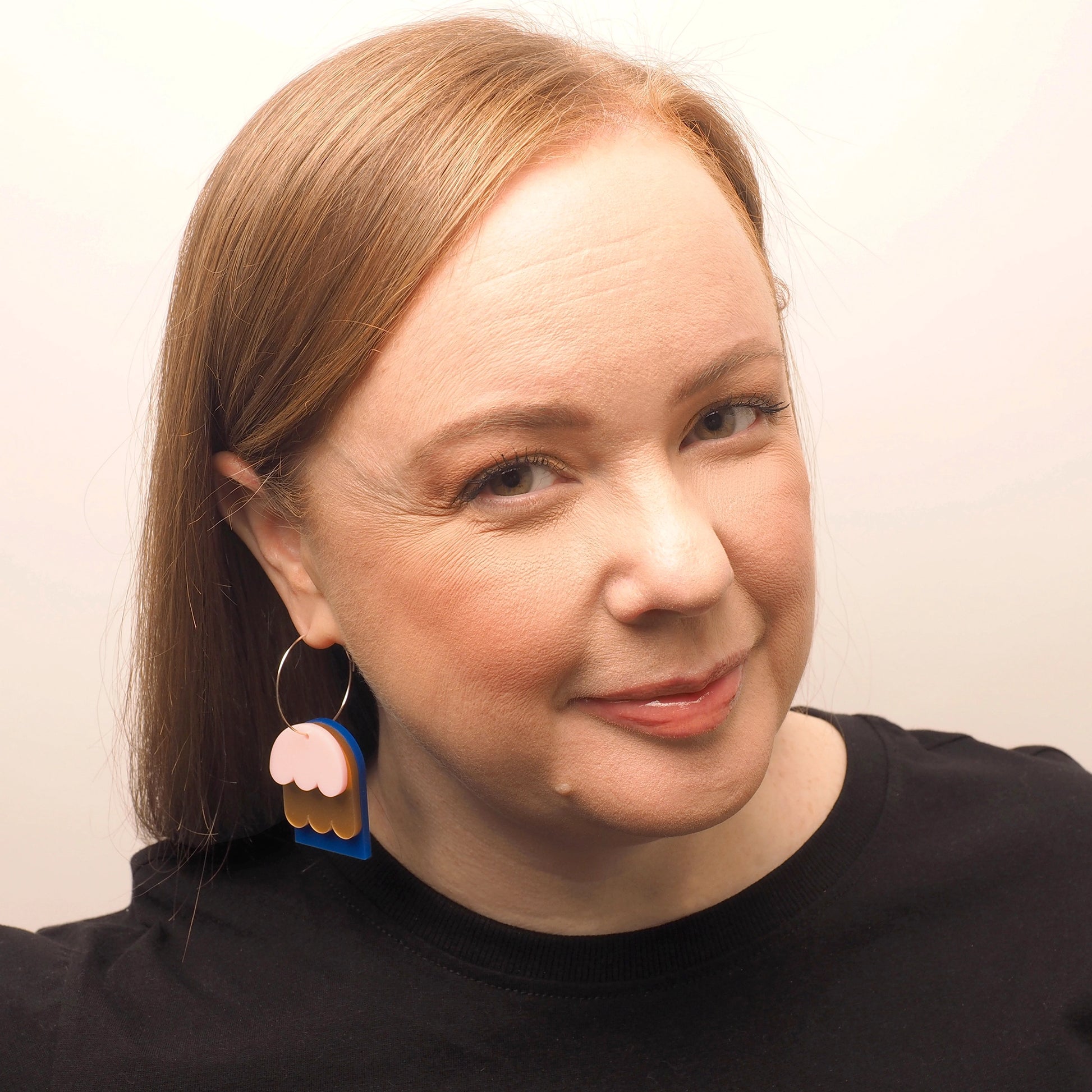 A curvy woman with straight red hair, wearing petal shaped earrings.These earrings have three layers, one is blue, one if pink and one is gold. They are hanging on a large hoop fixture.