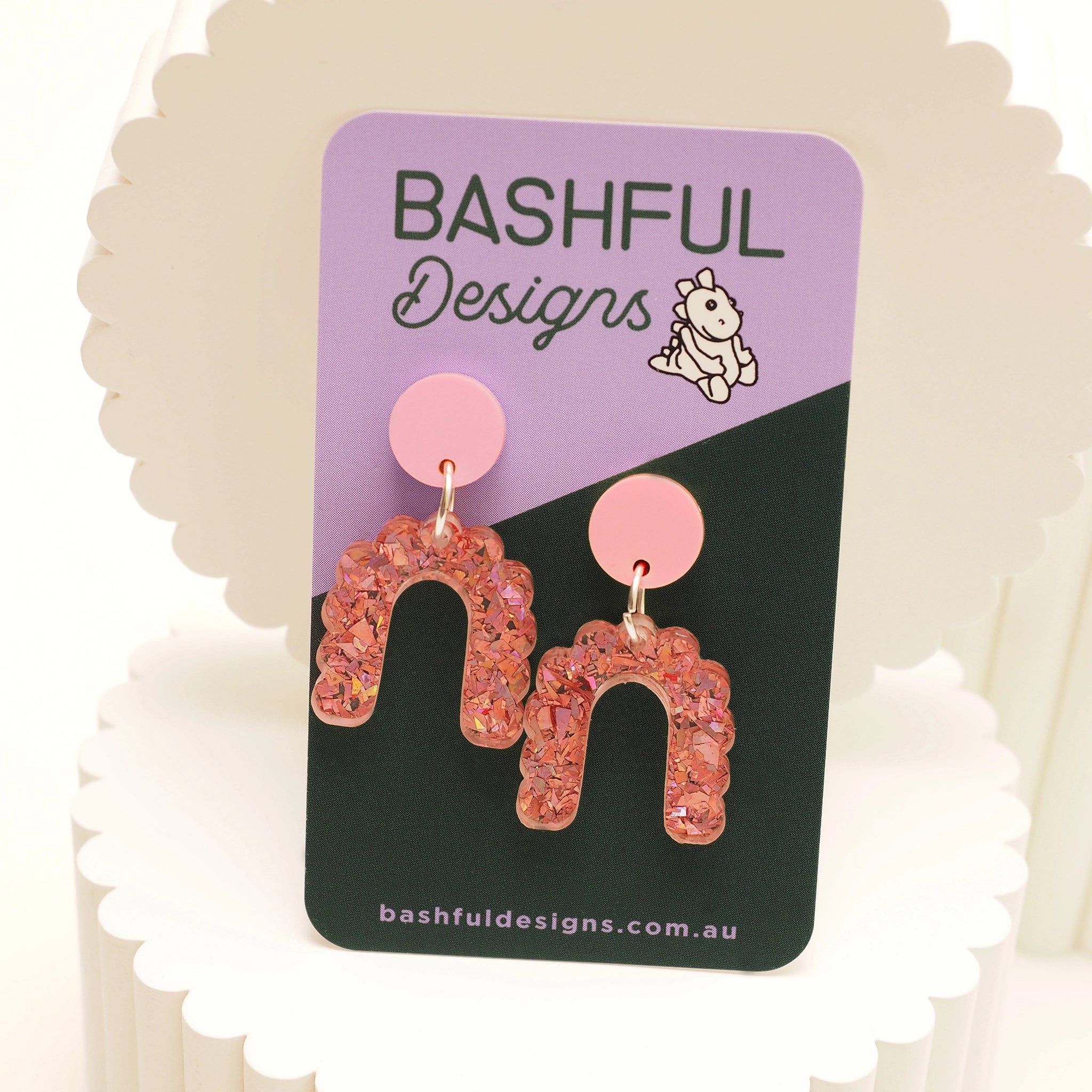 Pink glitter small dangle stud arch earrings, on a 'Bashful Designs' branded card.