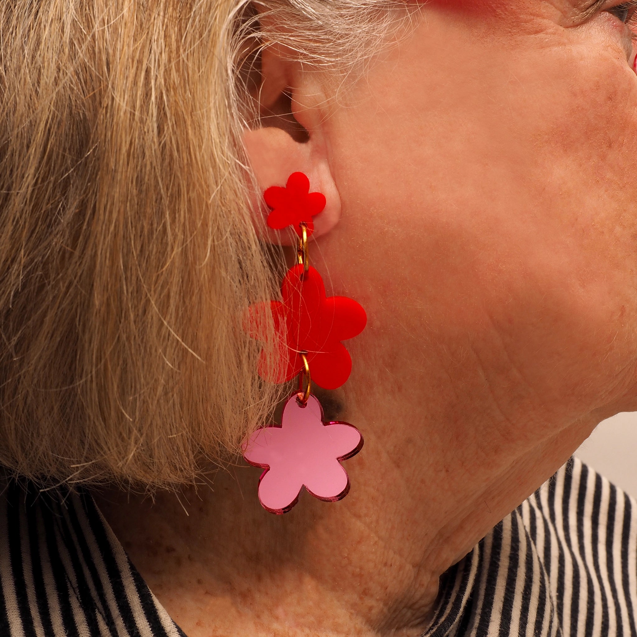 A person with grey and blonde hair, wearing flower shaped acrylic earrings. Two of the flower shapes are red acrylic and at the bottom of the earrings is a third flower, that is pink mirrored acrylic.