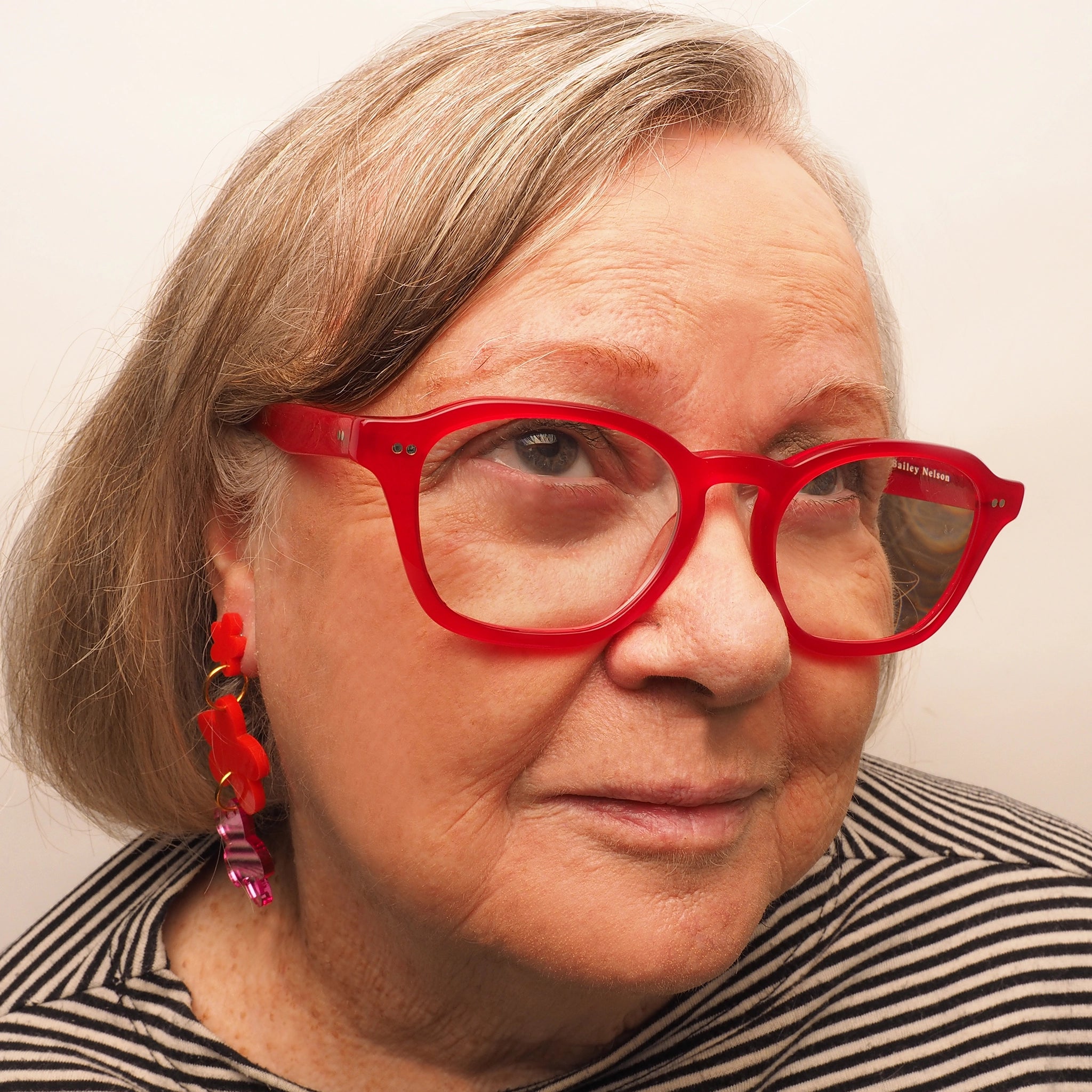 A woman in her 70s with grey and blonde hair, wearing red glasses and a pair of acrylic earrings in the shape of three flowers. The first tier is a small, red flower. The second tier is a medium-size red flower. The third tier is a medium-size, pink mirrored flower.