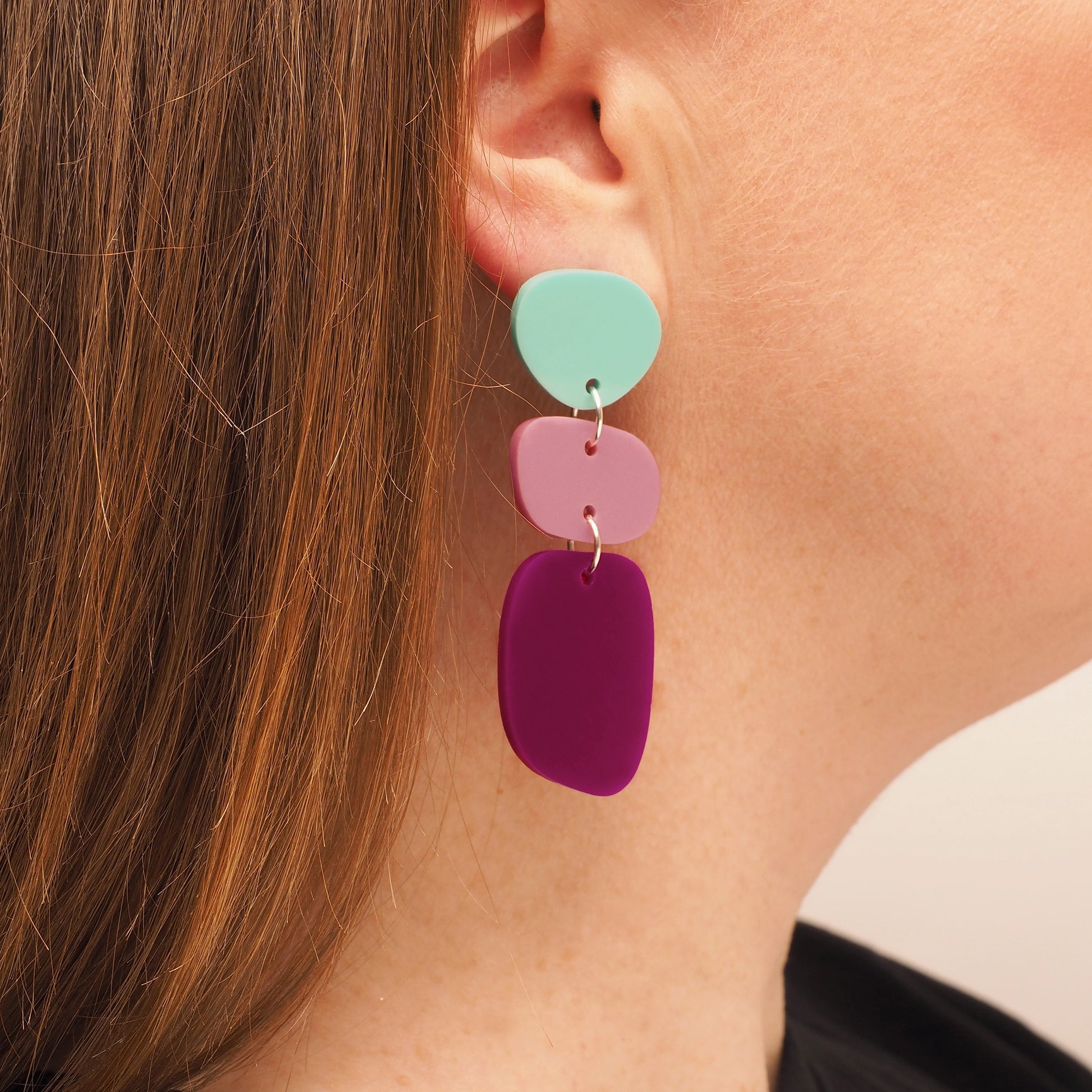 A person with straight red hair wearing pebble-shaped acrylic earrings. The earrings consist of three pebble shapes; the first is a light blue, the second is pink, and the third is a dark magenta.