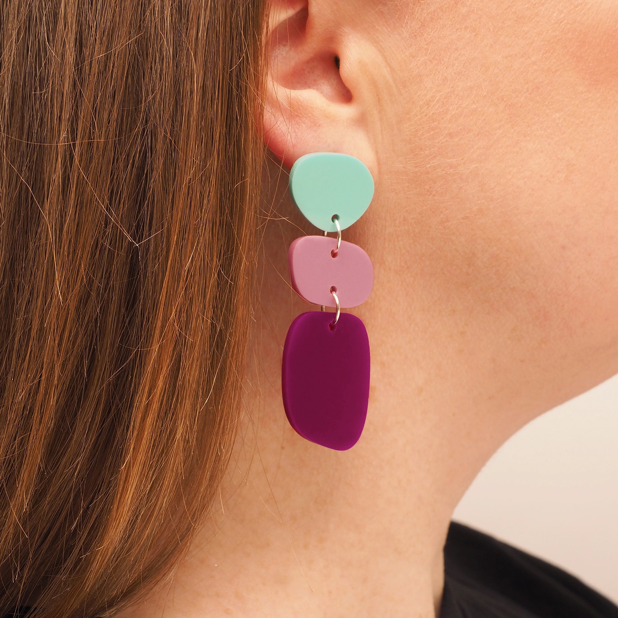 A person with straight red hair wearing pebble-shaped acrylic earrings. The earrings consist of three pebble shapes; the first is a light blue, the second is pink, and the third is a dark magenta.