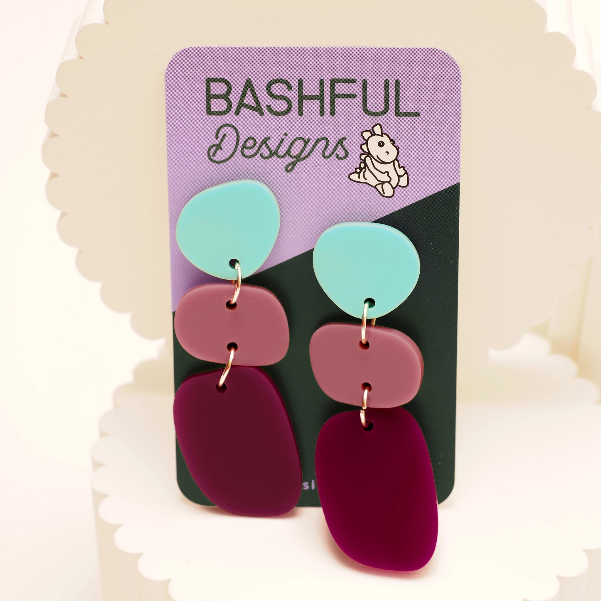 A pair of pebble-shaped acrylic dangle earrings, shown on a 'Bashful Design' product card. The earrings consist of three pebble shapes; the first is a light blue, the second pink and the third a dark magenta.