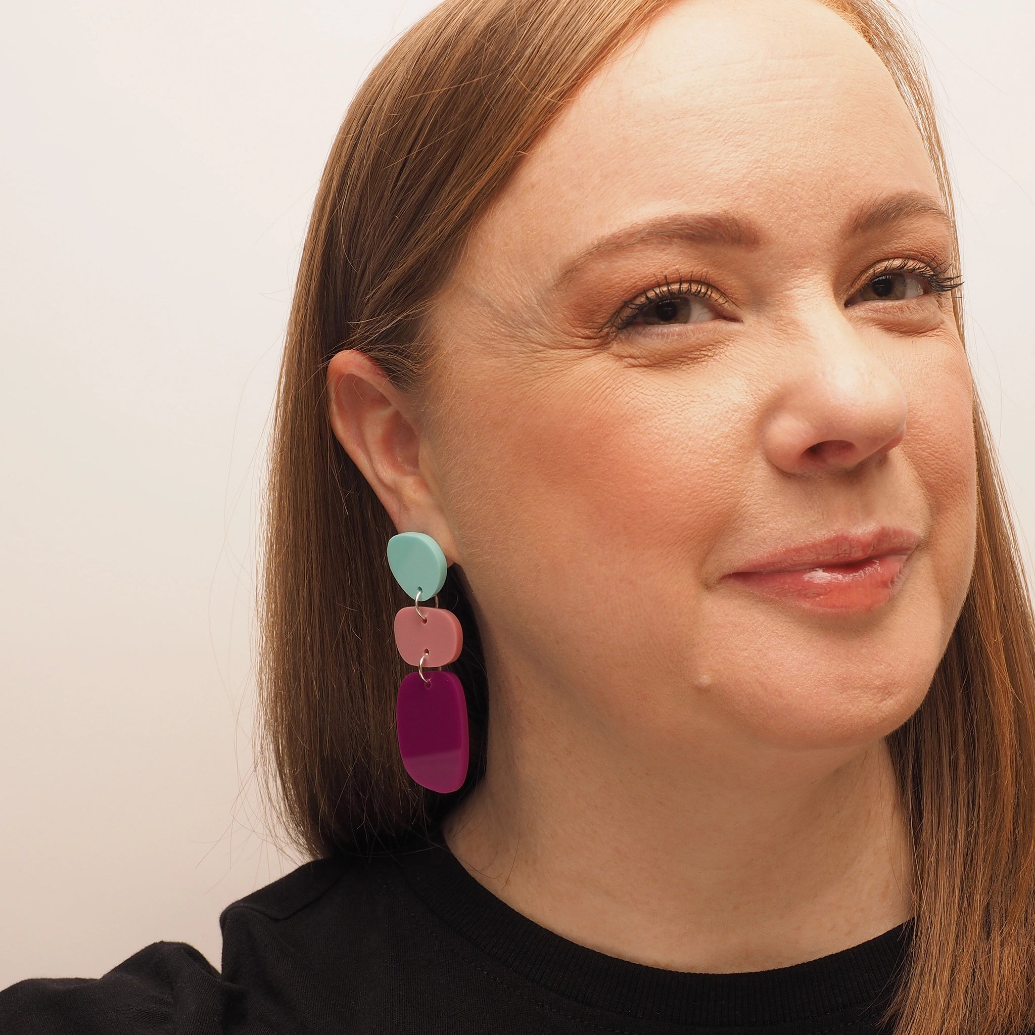 A curvy woman with red hair, wearing pebble-shaped acrylic earrings. There are three pebble shapes; the first is a light blue, the second is pink, and the third is dark magenta.
