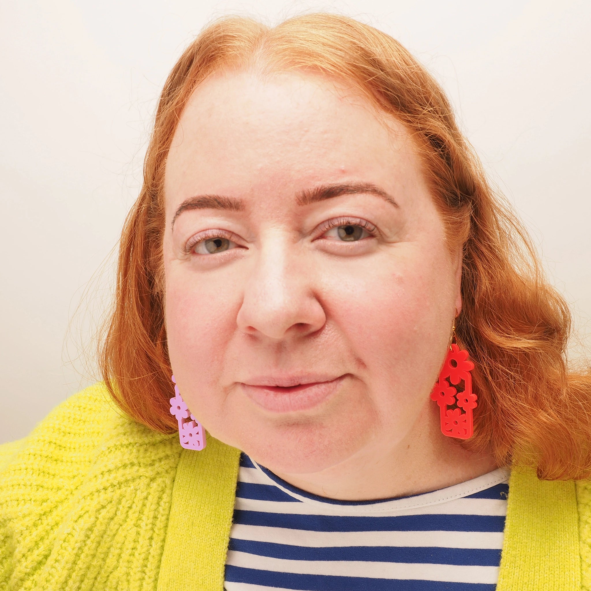 A curvy woman with curly red hair wearing lilac pink and bright red acrylic floral dangle earrings.