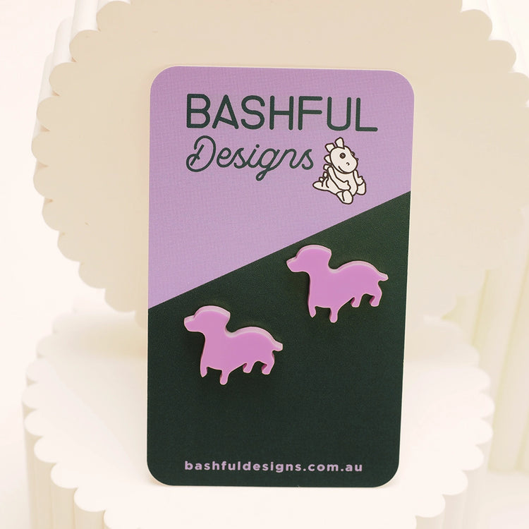 A close-up of pink sausage dog studs on a branded, purple and green earrings card.