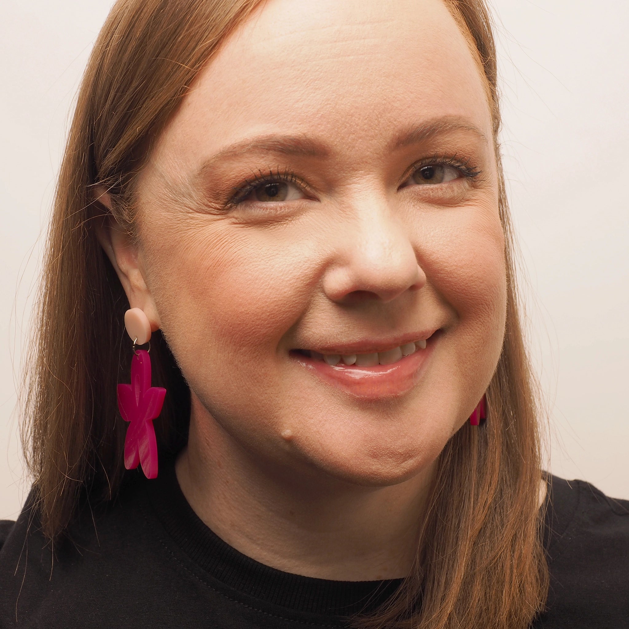 A woman with straight red hair wearing bright pink acrylic earrings in the shape of a wild flower.