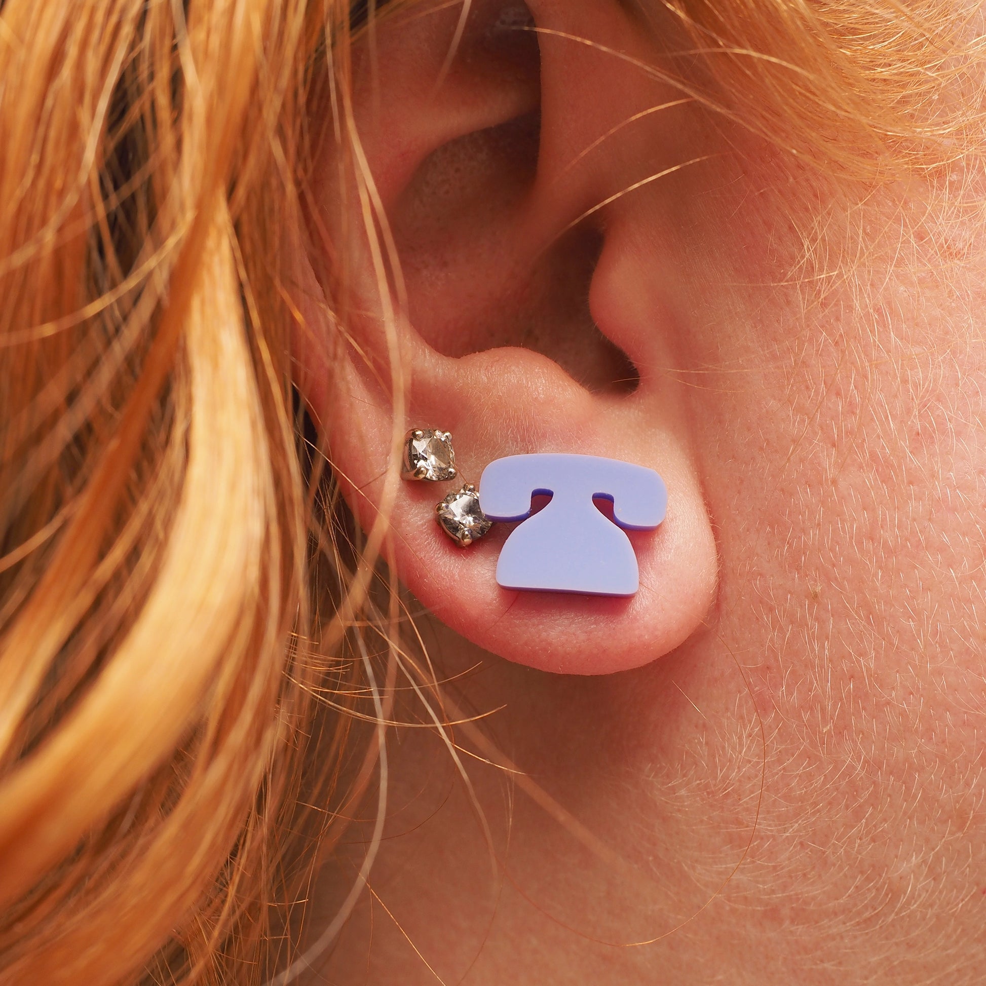 A person with red curly hair, wearing light purple earrings in the shape of a retro telephone.