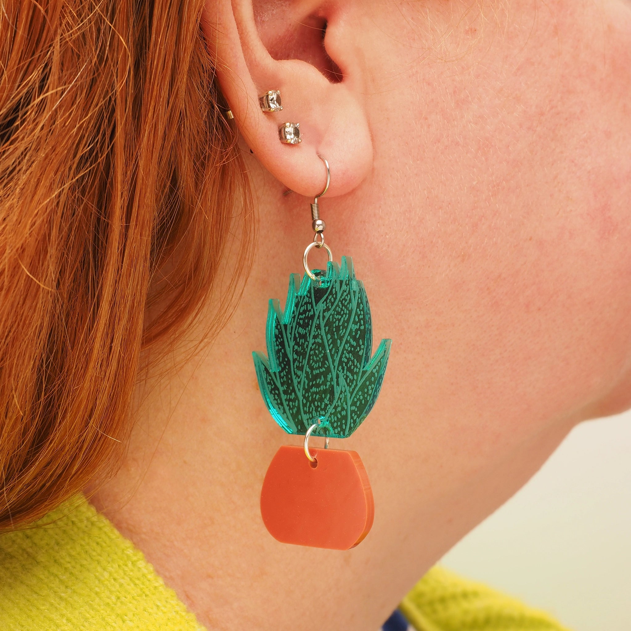 A person with curly red hair, wearing acrylic earrings in the shape of a green, spiky agave plant, in a terracotta plant pot.