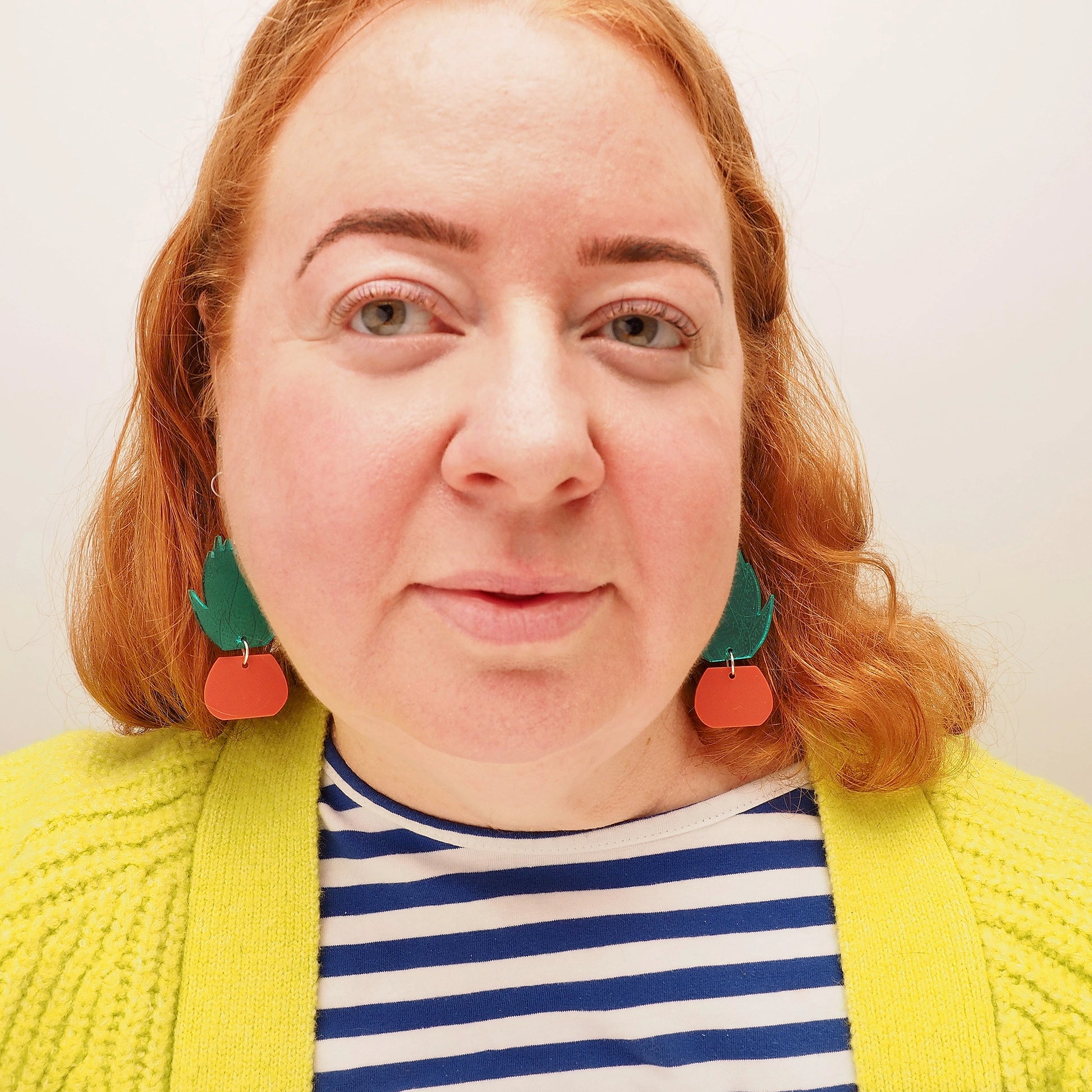A curvy woman with red hair, wearing acrylic earrings in the shape of a green, spiky agave plant in a terracotta plant pot.