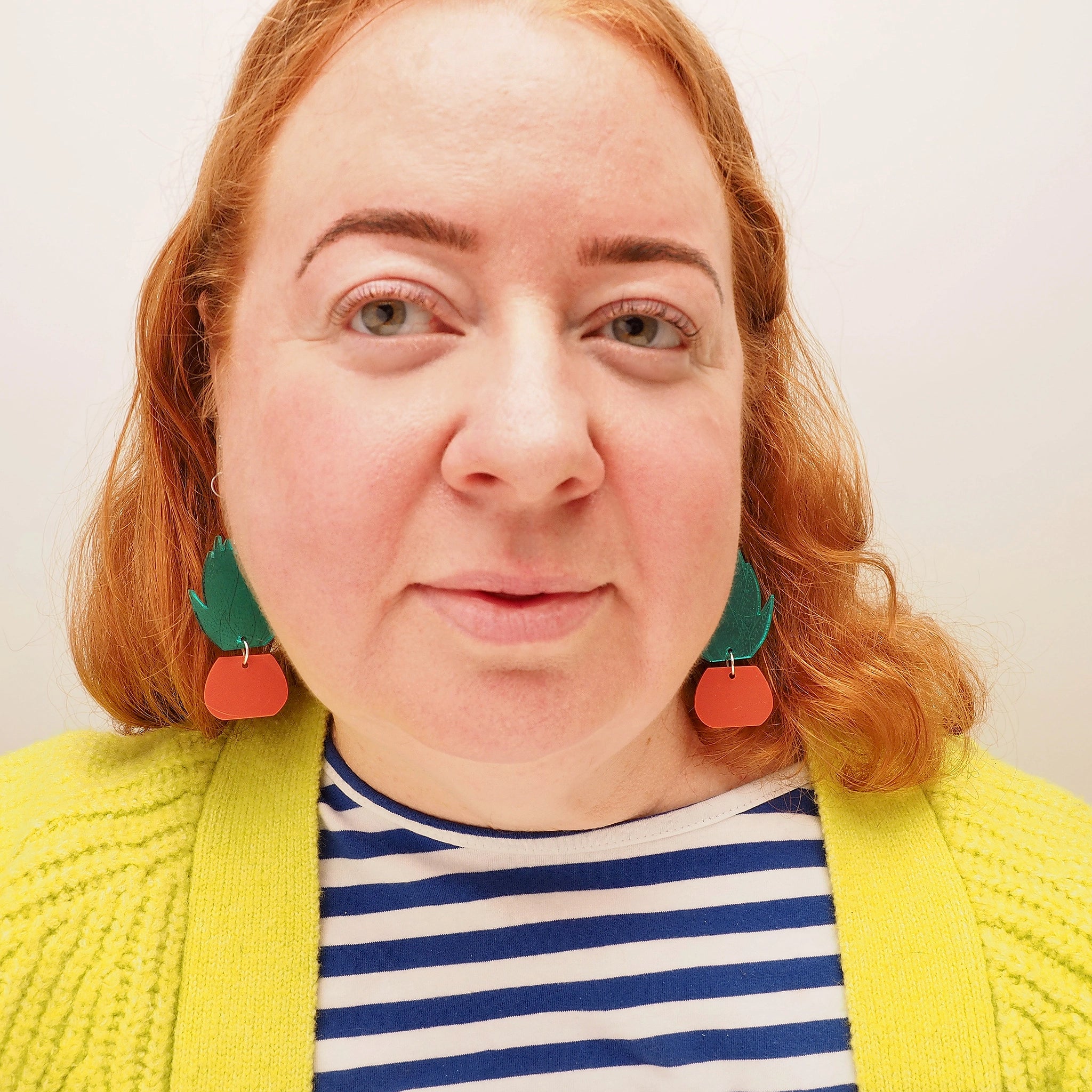 A curvy woman with red hair, wearing acrylic earrings in the shape of a green, spiky agave plant in a terracotta plant pot.