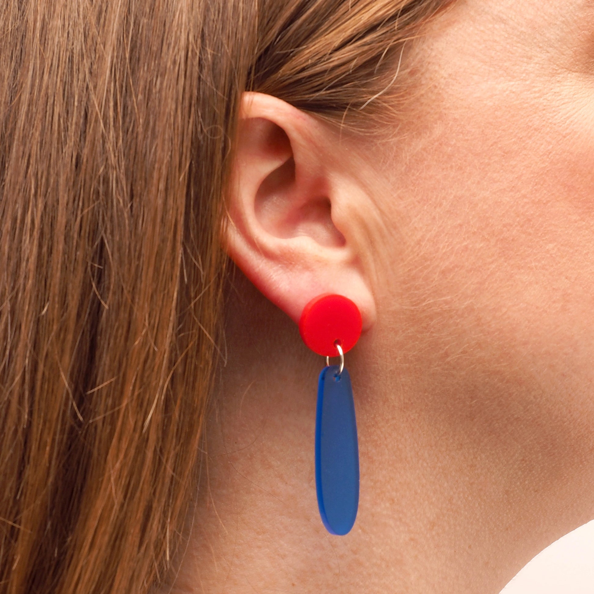 A person with straight red hair wearing a pair of red and blue earrings. They begin with a round red topper and follow with a long droplet shape in opaque navy.