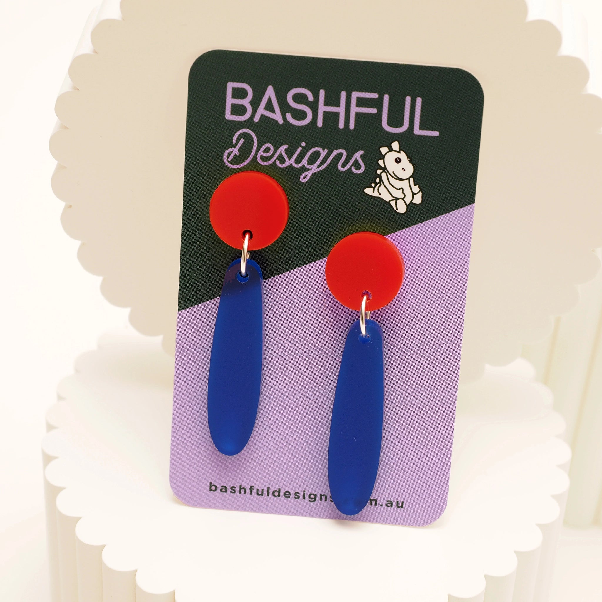 Acrylic earrings that begin with a red earring topper and follow with a long droplet shape in opaque navy. They are shown on a 'Bashful Designs' earring card.