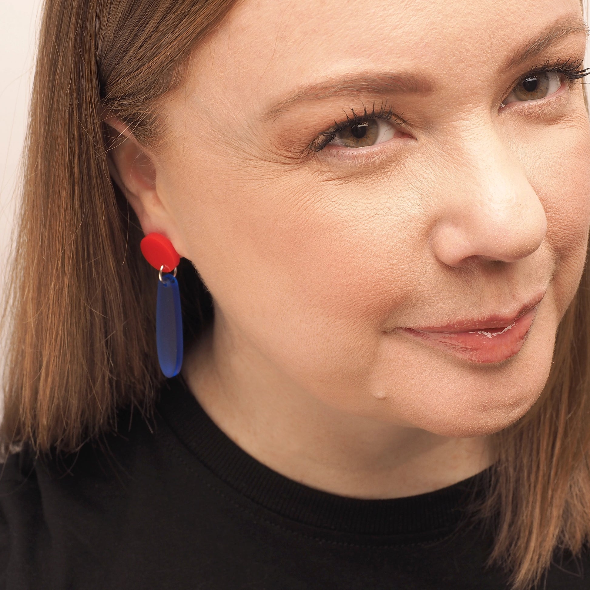 A woman with straight red hair wearing a pair of red and blue earrings. They begin with a round red topper and follow with a long droplet shape in opaque navy.