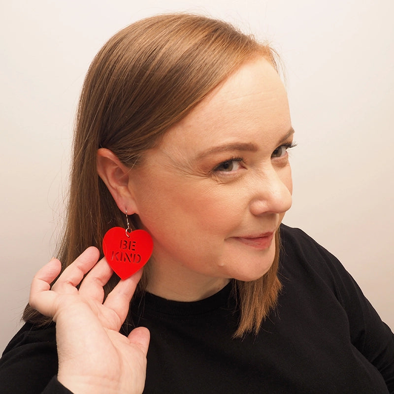 A curvy woman with straight, red hair smiling and wearing large, red heart earrings. The hearts earrings have the words 'Be Kind' laser-cut into the acrylic.