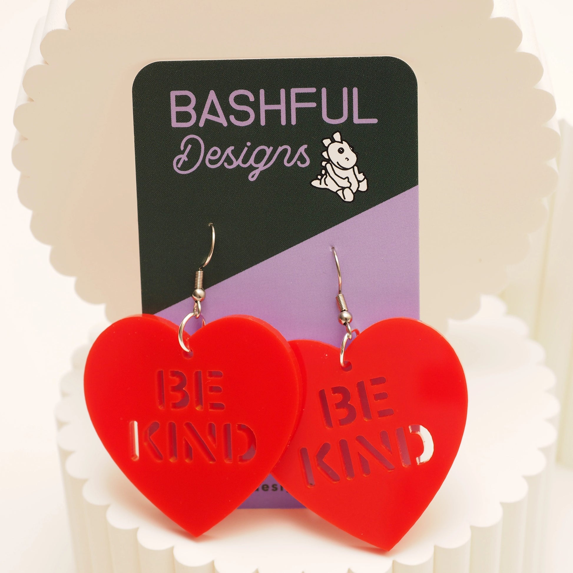 Solid red acrylic earrings in the shape of a heart with cut out wording that says "Be Kind". These earrings are shown on a 'Bashful Designs' product card.