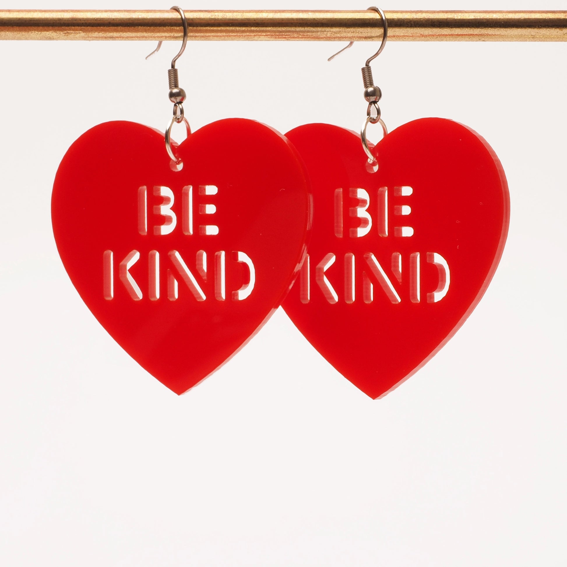 Solid red acrylic earrings in the shape of a heart with cut out wording that says "Be Kind". These earrings are shown hanging from a gold earring stand.