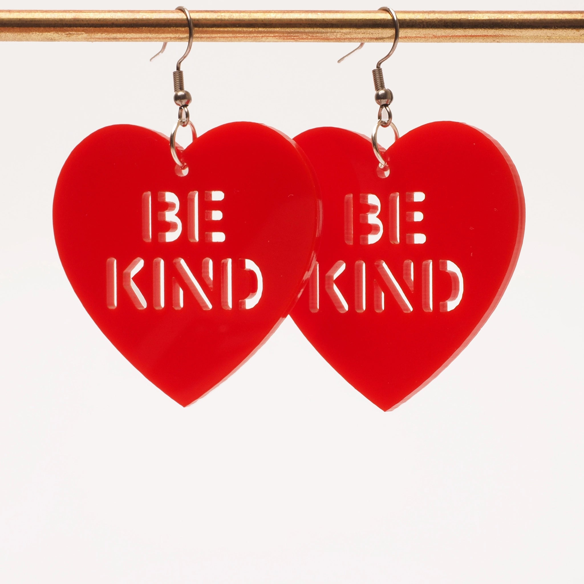 Solid red acrylic earrings in the shape of a heart with cut out wording that says "Be Kind". These earrings are shown hanging from a gold earring stand.