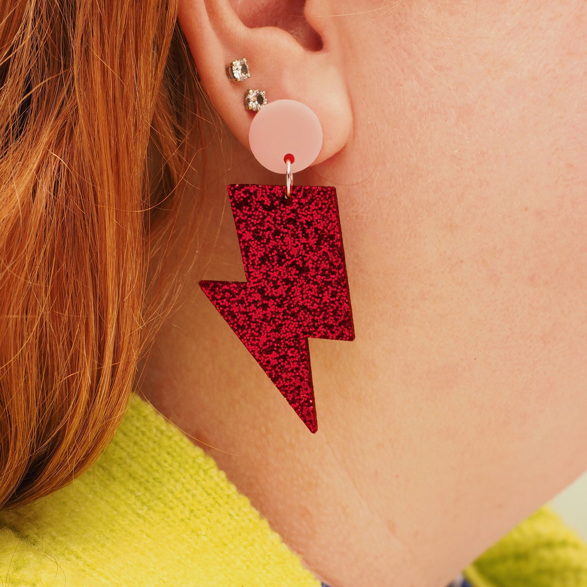 A person with red hair wearing a red glitter acrylic lightning bolt earring with a small, round, light pink earring topper.