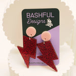 Red glitter acrylic earrings in the shape of a lightning bolt. They also feature a light pink earring topper, and are shown on a 'Bashful Designs' earring card.