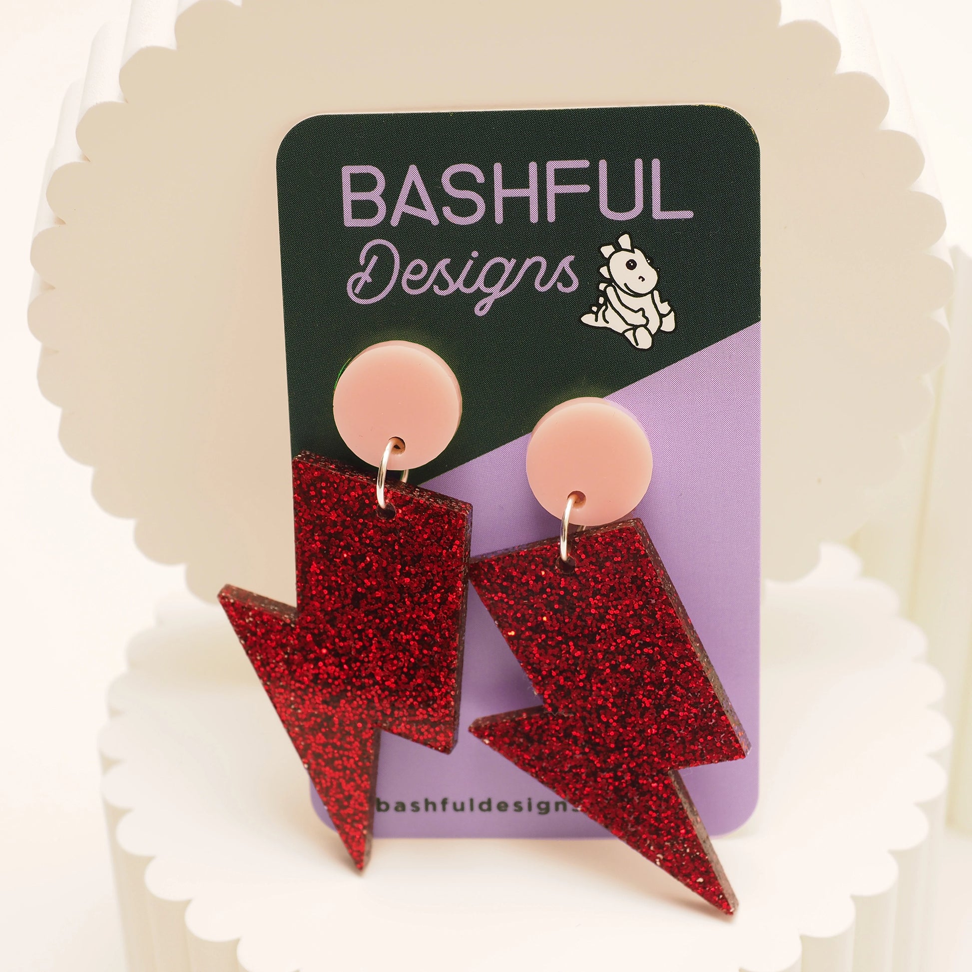 Red glitter acrylic earrings in the shape of a lightning bolt. They also feature a light pink earring topper, and are shown on a 'Bashful Designs' earring card.