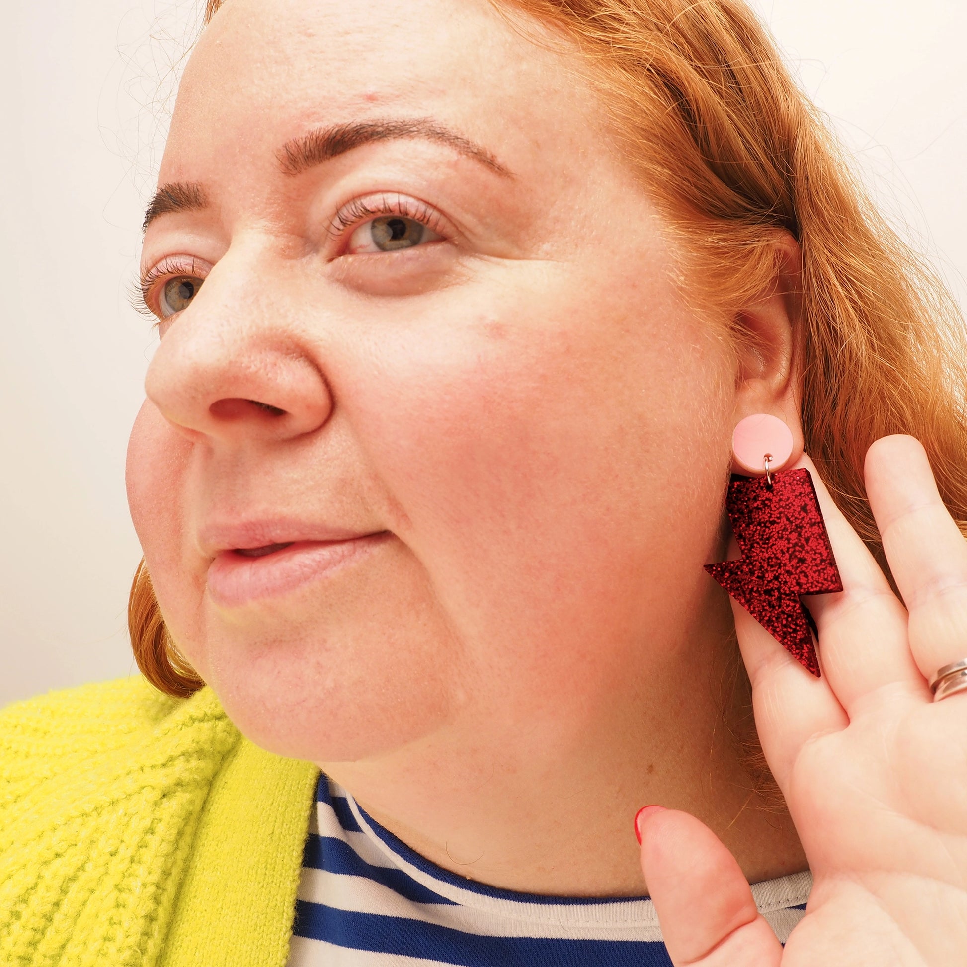 A curvy woman with red hair wearing a pair of red glitter acrylic earrings in the shape of a lightning bolt. They also feature a small, round, light pink earring topper.