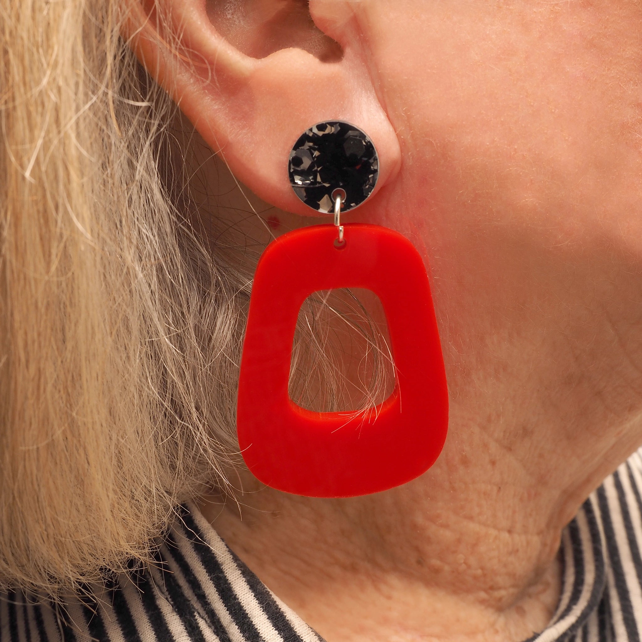 A woman with grey and blonde hair wearing orange-red oblong shaped earrings with a black and silver glitter topper.