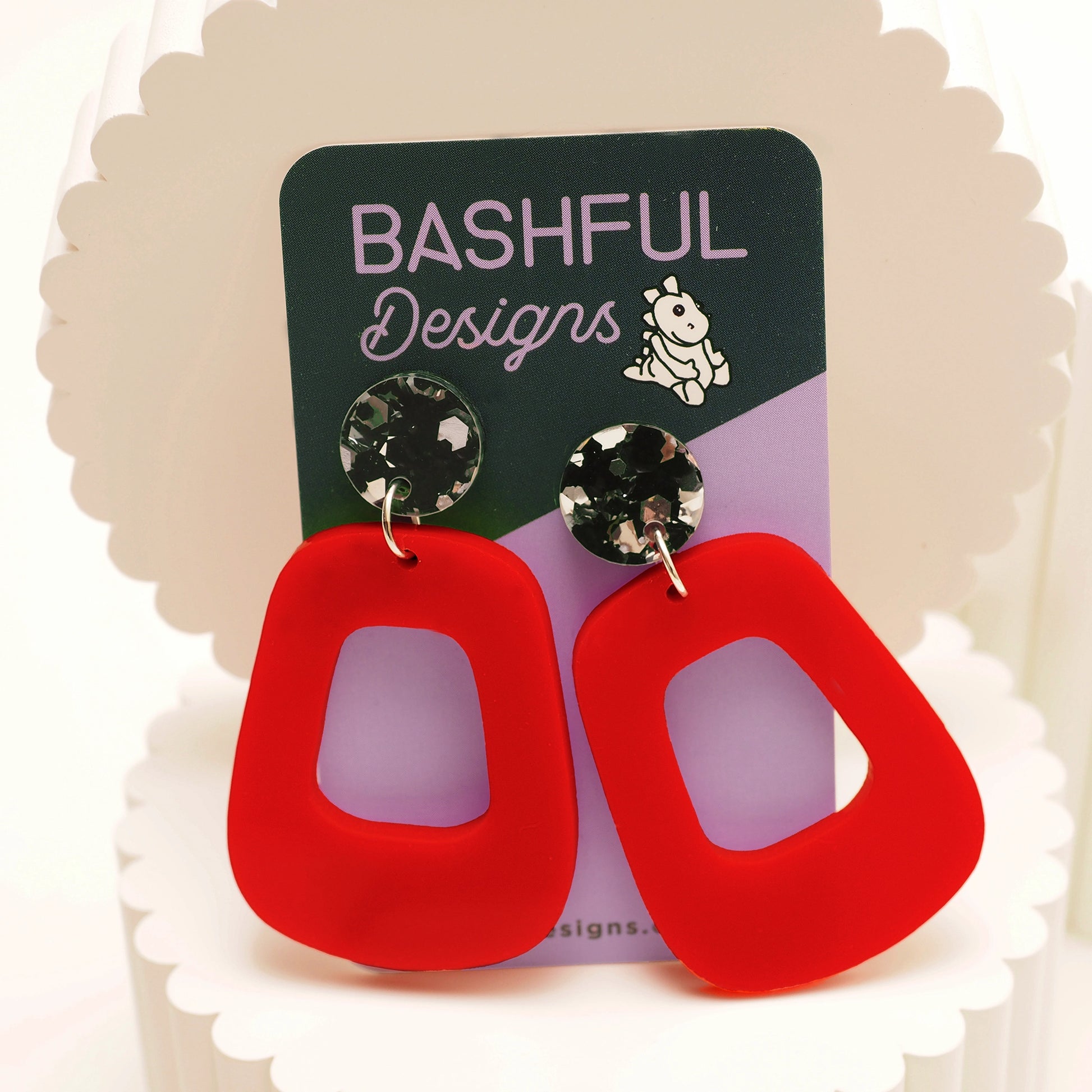 Orange-red oblong shaped earrings with a black and silver glittery earring topper. The earrings are pictured on a 'Bashful Designs' card.