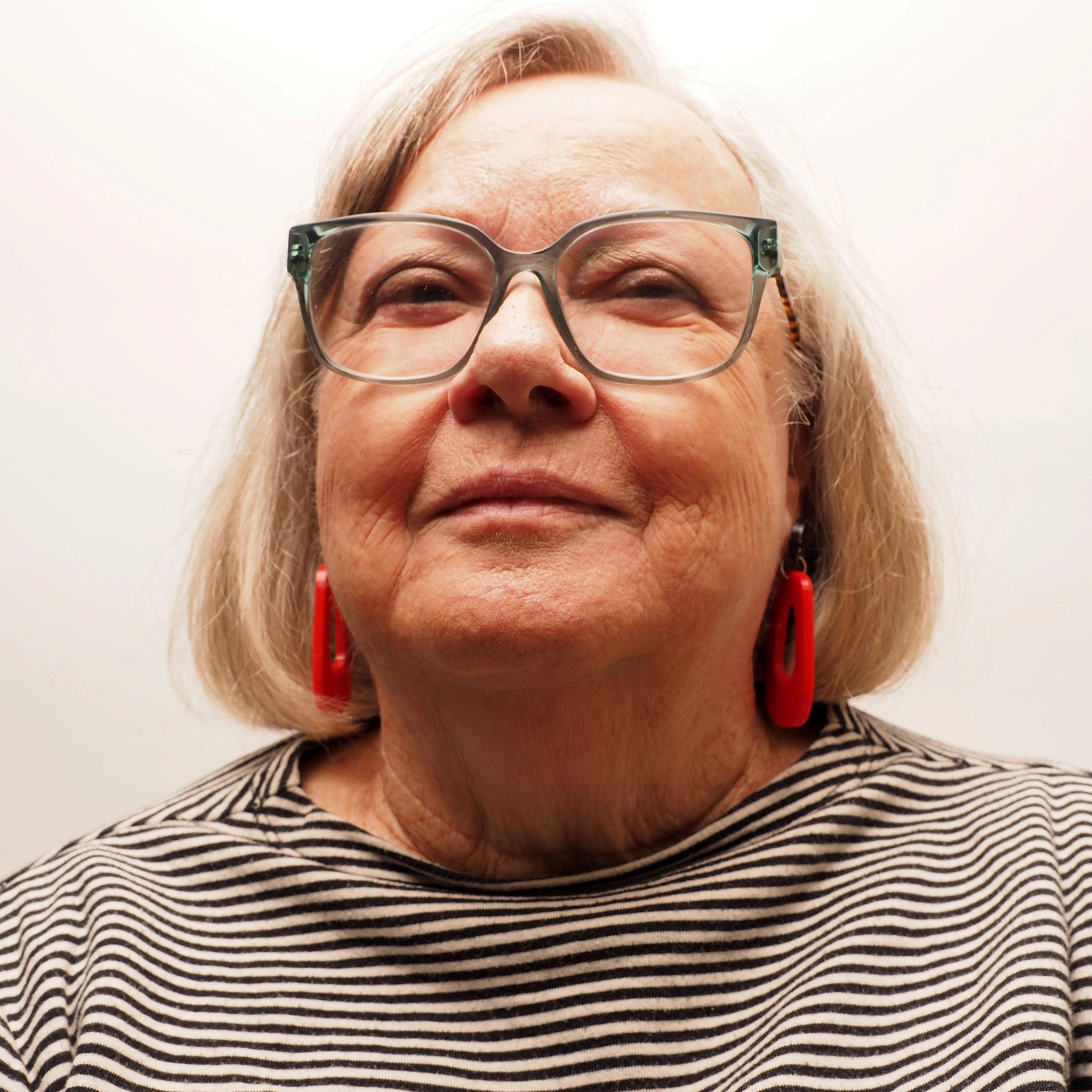 A woman in her 70's with grey and blonde hair wearing a pair of orange-red oblong shaped earrings. The earrings have a black and silver glittery topper.