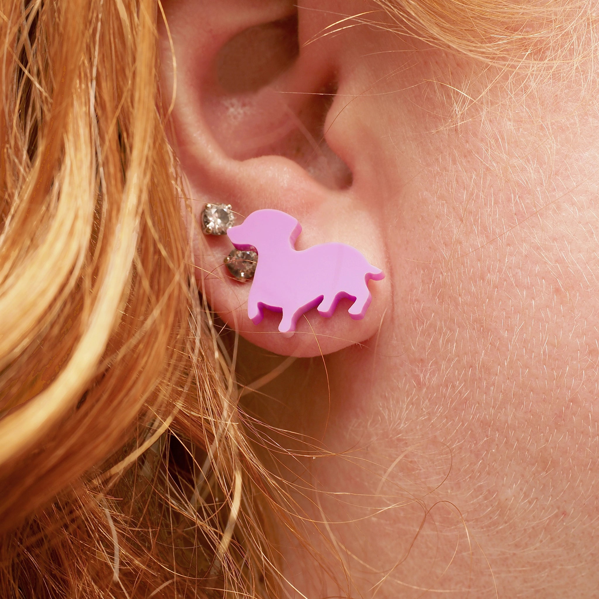 A woman with red and blonde hair wearing a pair of pink stud earrings in the shape of a sausage dog.