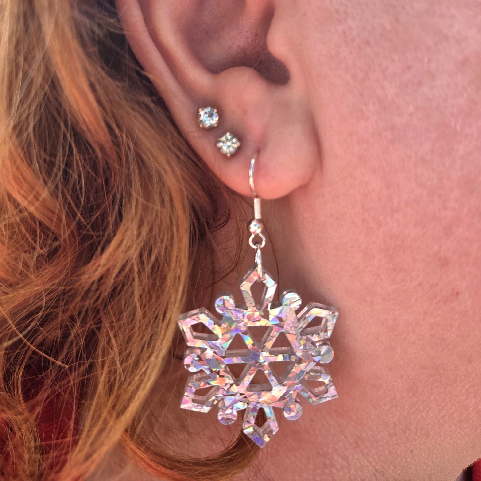 Earrings in the shape of a snowflake, made from silver holographic acrylic.   They are shown hanging in a person's ear.