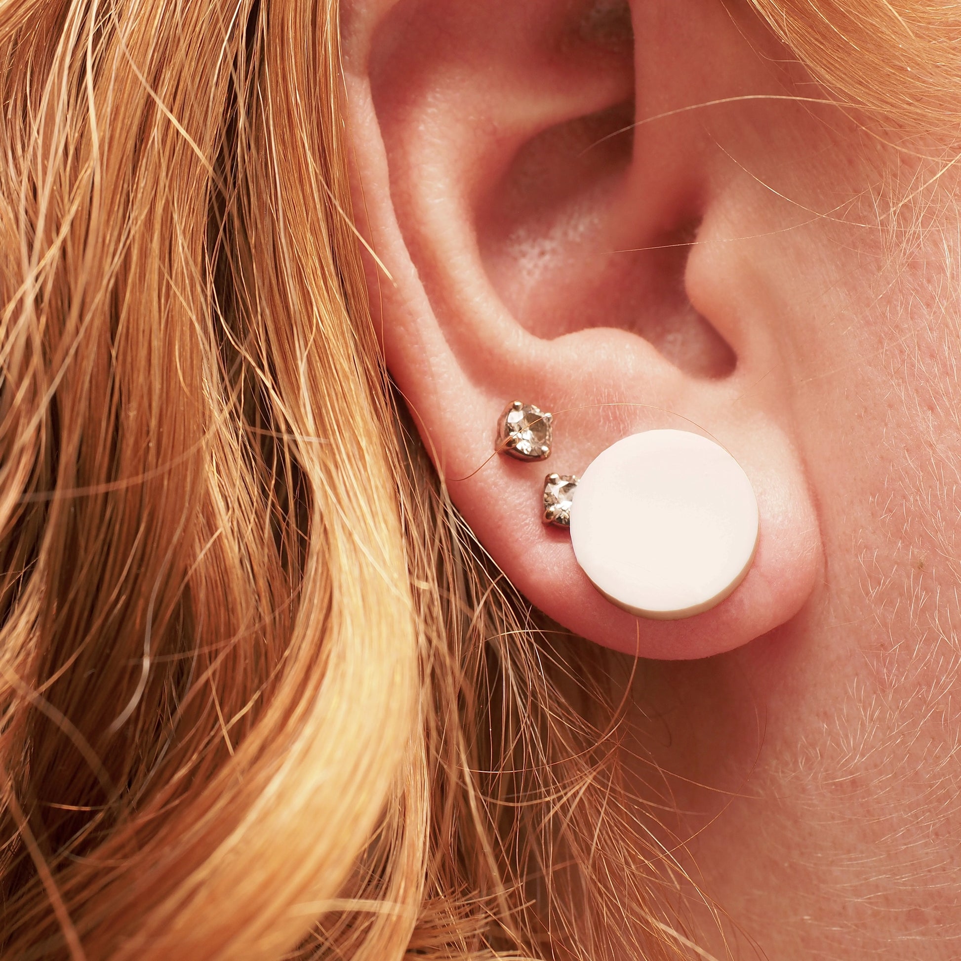 A person with red and blonde hair wearing white acrylic earrings in the shape of a dot.