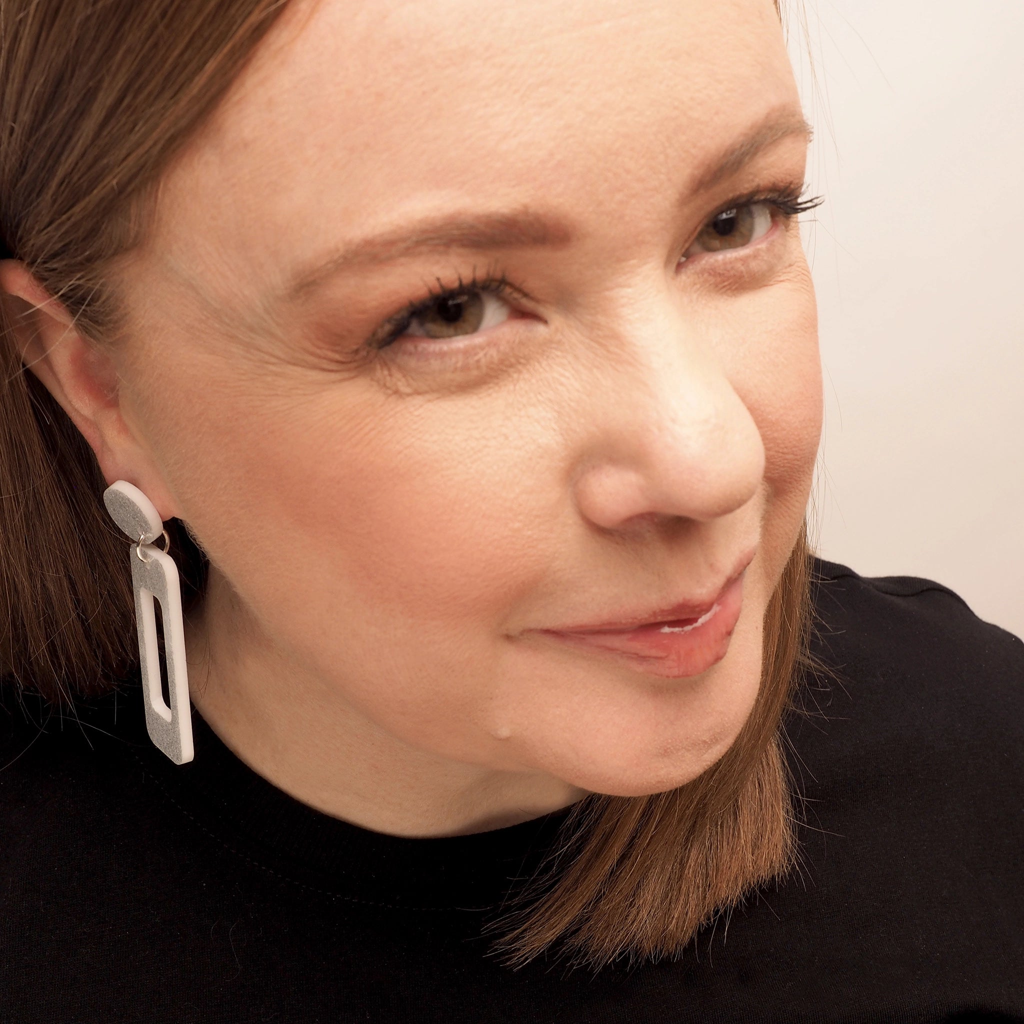 A woman with straight red hair wearing a pair of white glitter acrylic earrings in a rectangle shape. They also feature a small, round, white glitter earring topper.
