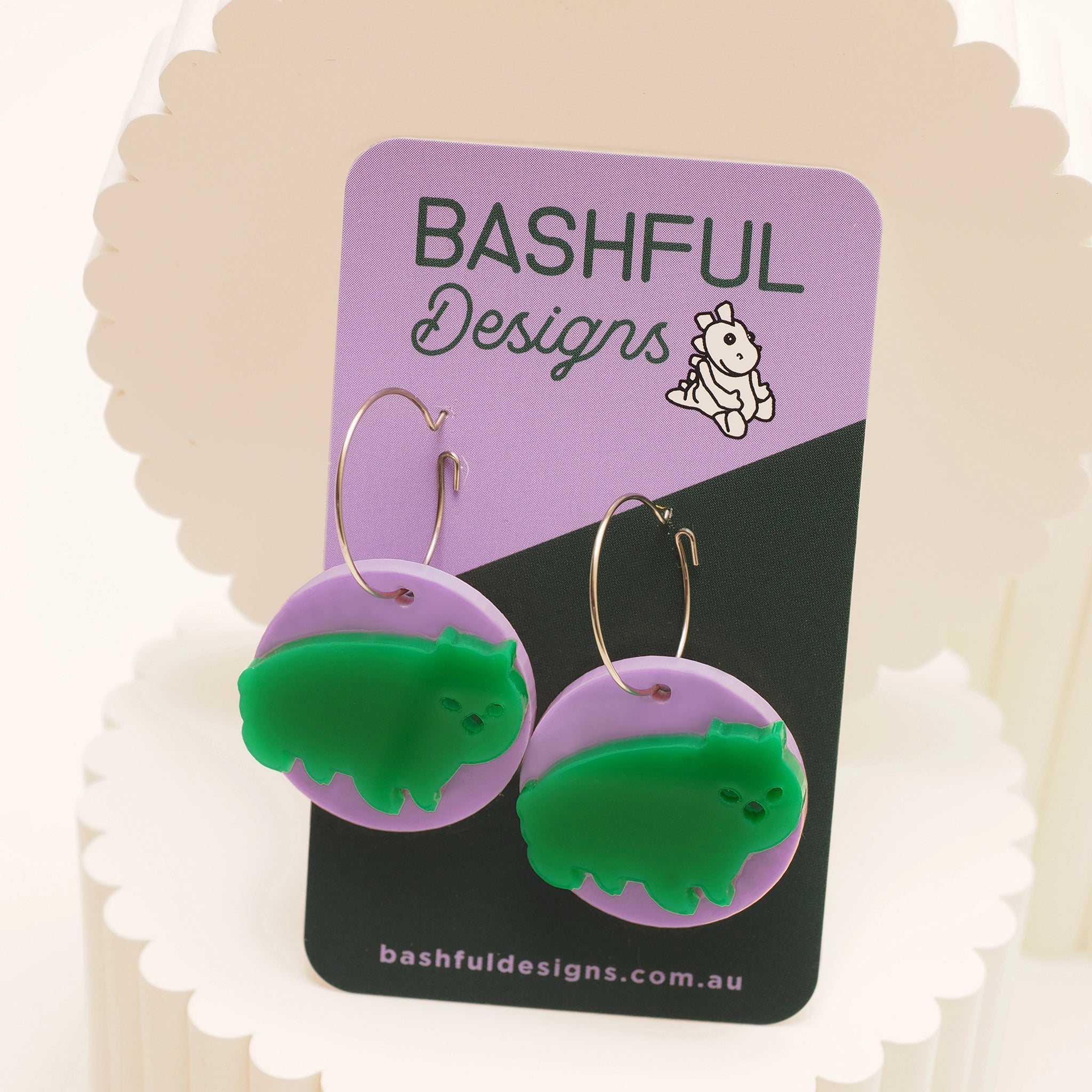 Green acrylic earrings shaped like a wombat stuck to a pink acrylic circle. These earrings are hanging from a medium sized silver hoop, and shown on a 'Bashful Designs' earring card.