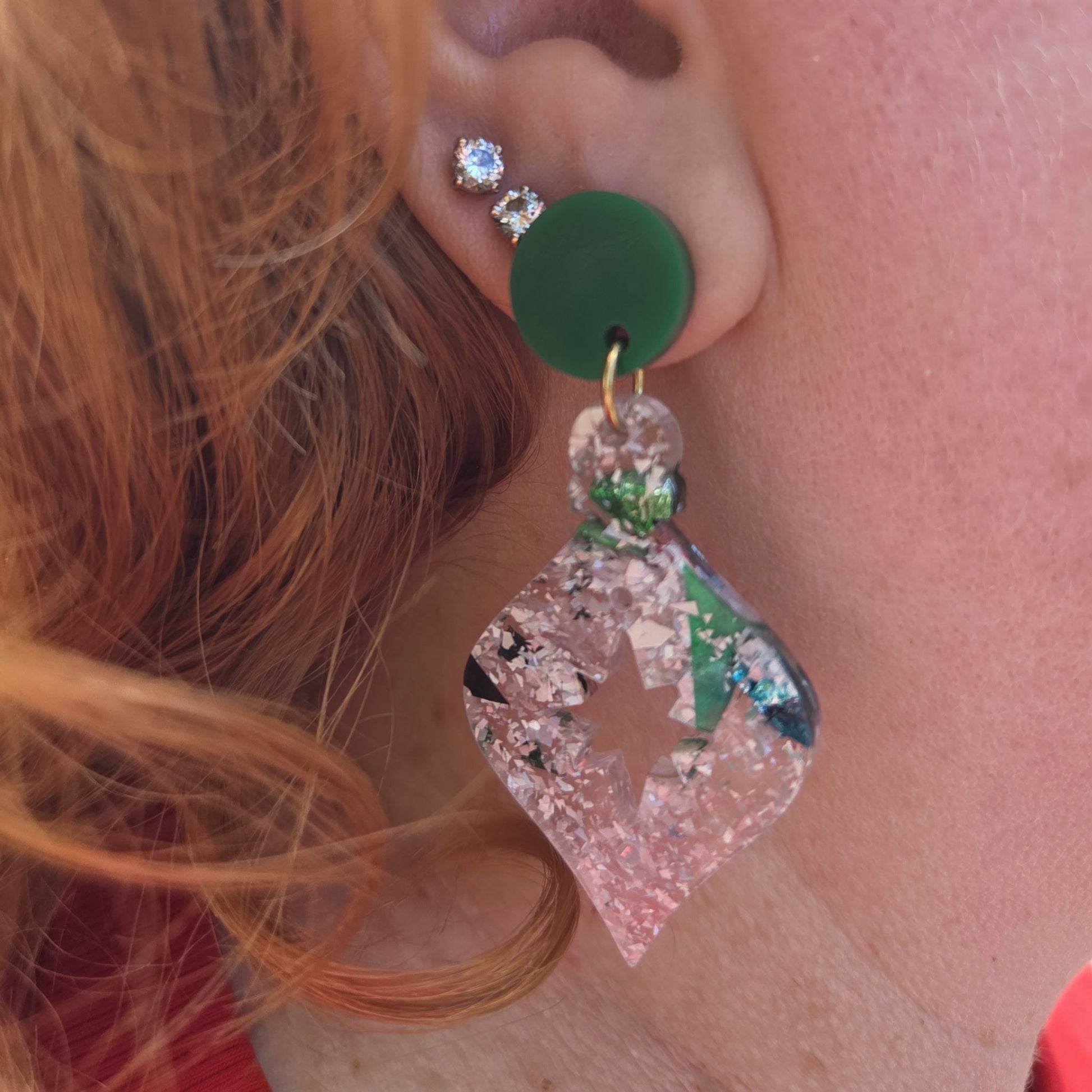 Earrings in the shape of a Christmas Tree bauble.  The earrings begin with a dark green acrylic topper, then flow into a bauble shape in silvery chunky glitter acrylic.  They are shown in a person's ear.