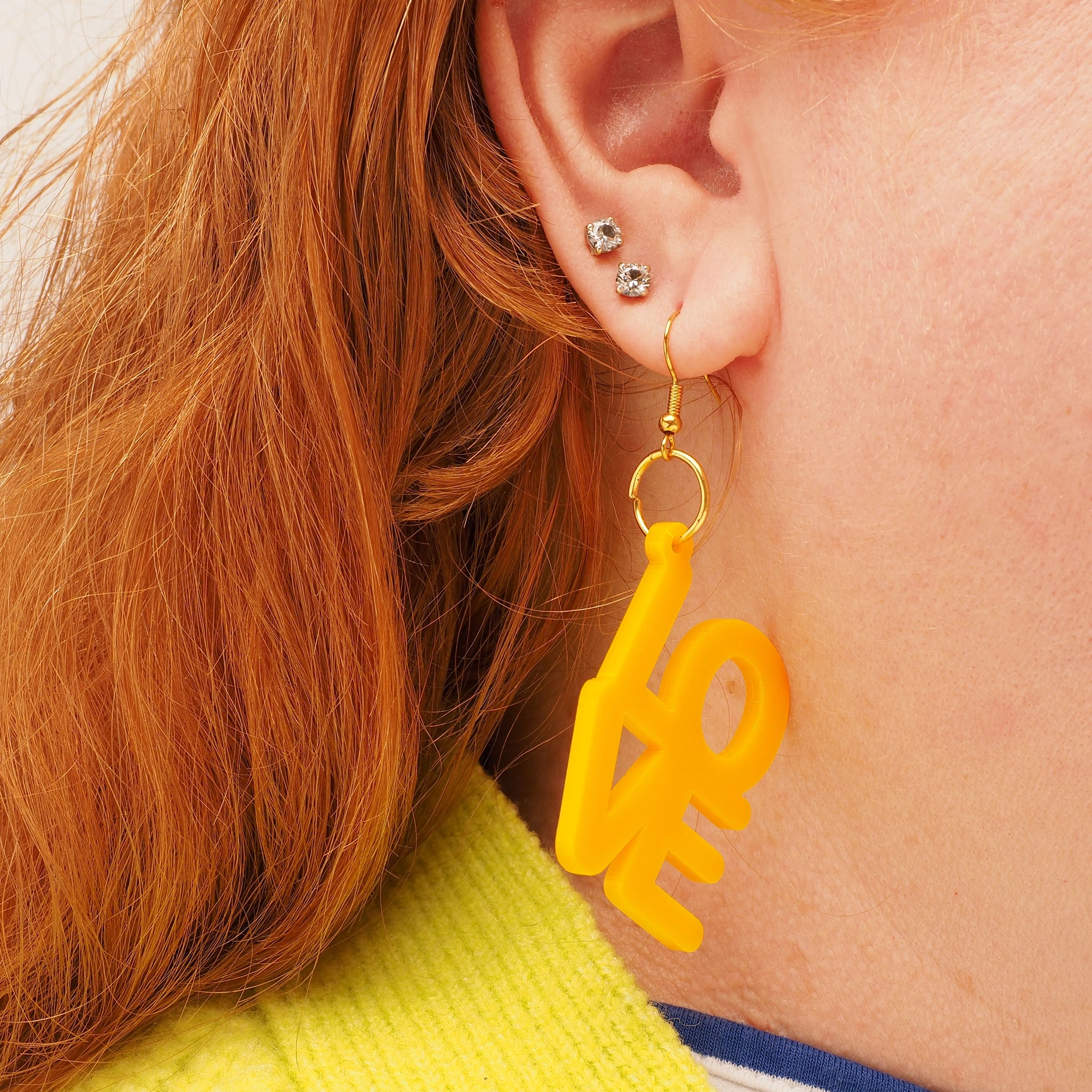 A person with long red hair wearing a yellow earring that spells out the word 'love'.