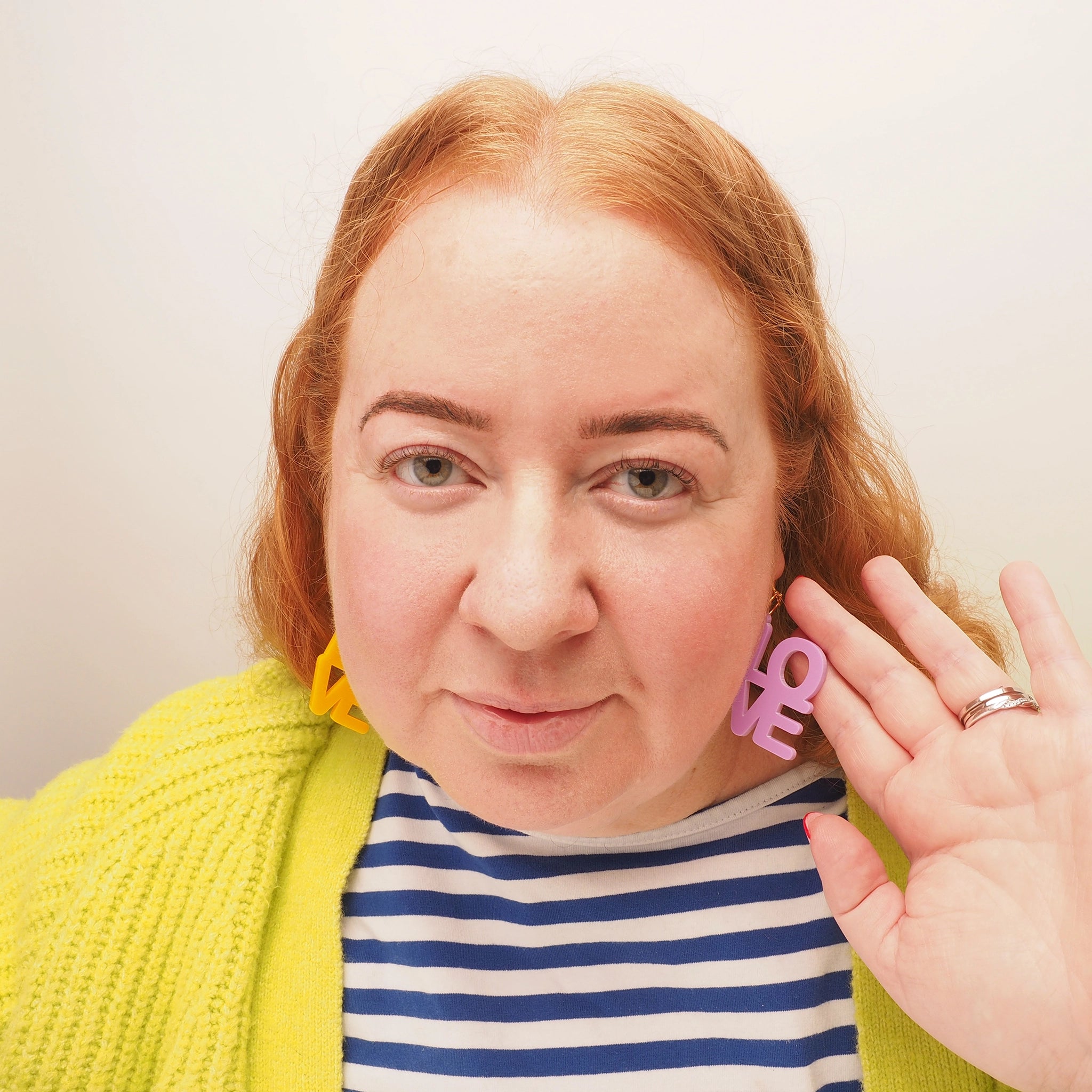 A woman with long red hair wearing acrylic earrings that spell out the word 'love'. One earring is in yellow, and the other earring is a purple-y pink.