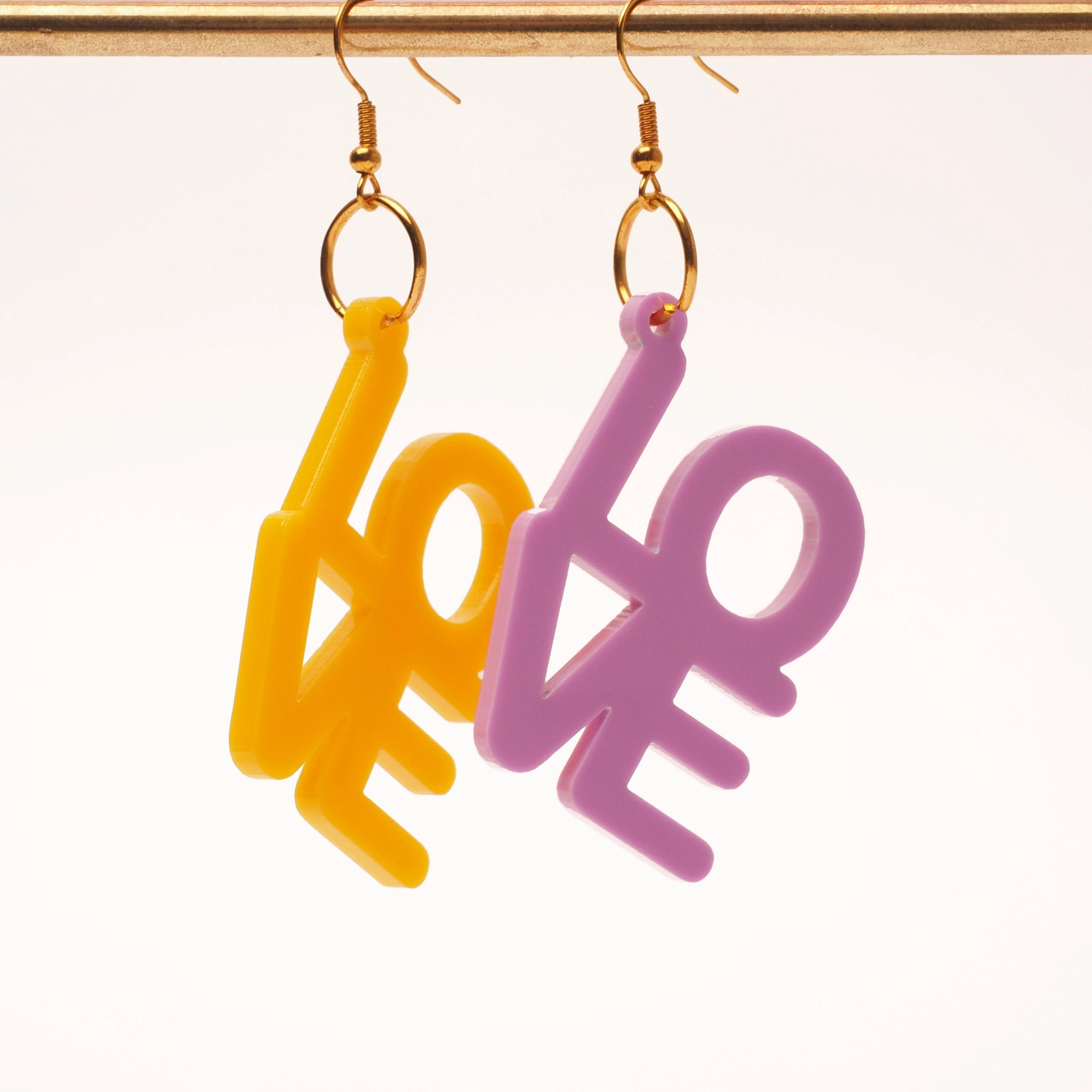 Acrylic earrings that spell out the word 'love'. One earring is in yellow, and the other earring is purple-y pink. These earrings are shown hanging on a gold earring stand.