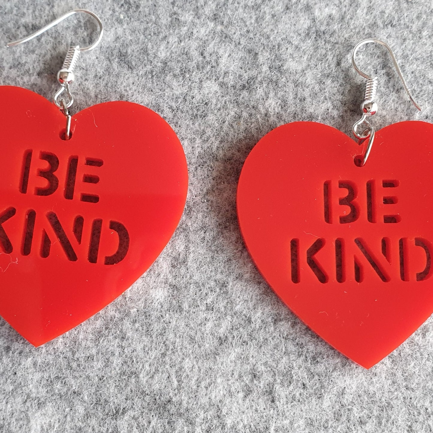 Red Be Kind Heart Earrings, facing front on to the viewer, on a grey felt background. These earrings are made from a red solid acrylic, with the words "Be Kind" cut out.