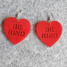 Load image into Gallery viewer, Red Be Kind Heart Earrings, facing front on to the viewer, on a grey felt background. These earrings are made from a red solid acrylic, with the words &quot;Be Kind&quot; cut out. The photo is a pulled-back view.
