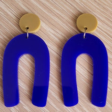 Load image into Gallery viewer, The Blue Wishbone earrings start with an olive green solid acrylic circle stud, and then continue to an upside-down U shape in an opaque blue acrylic.  The earrings are sitting on a wooden table.