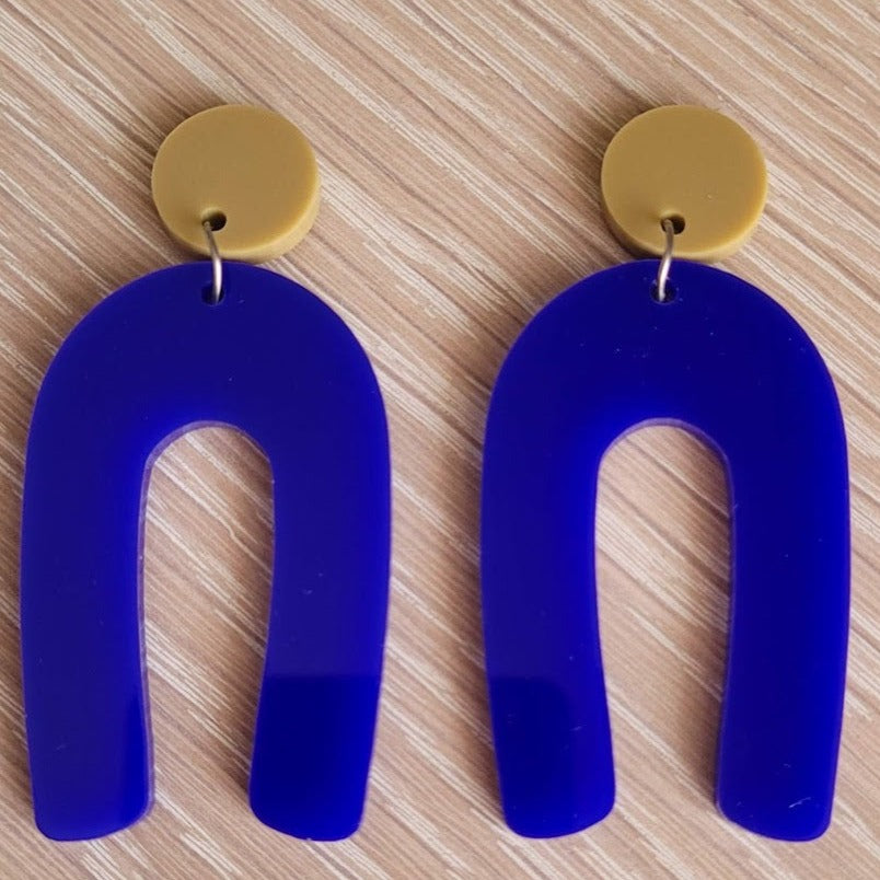 The Blue Wishbone earrings start with an olive green solid acrylic circle stud, and then continue to an upside-down U shape in an opaque blue acrylic.  The earrings are sitting on a wooden table.