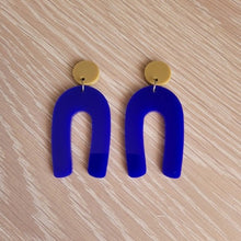 Load image into Gallery viewer, The Blue Wishbone earrings start with an olive green solid acrylic circle stud, and then continue to an upside-down U shape in an opaque blue acrylic. The earrings are sitting on a wooden table.